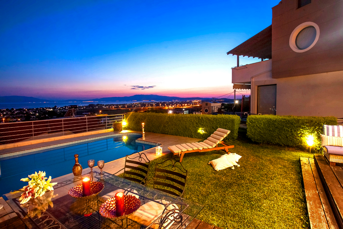 Outdoor patio with a pool, stylish outdoor lighting, lounge chairs, and a dining table with candles, overlooking a city and coastline at sunset.