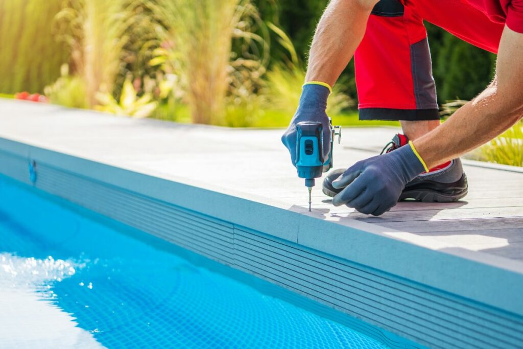 Worker using a drill to perform seasonal pool maintenance on the edge of a swimming pool.