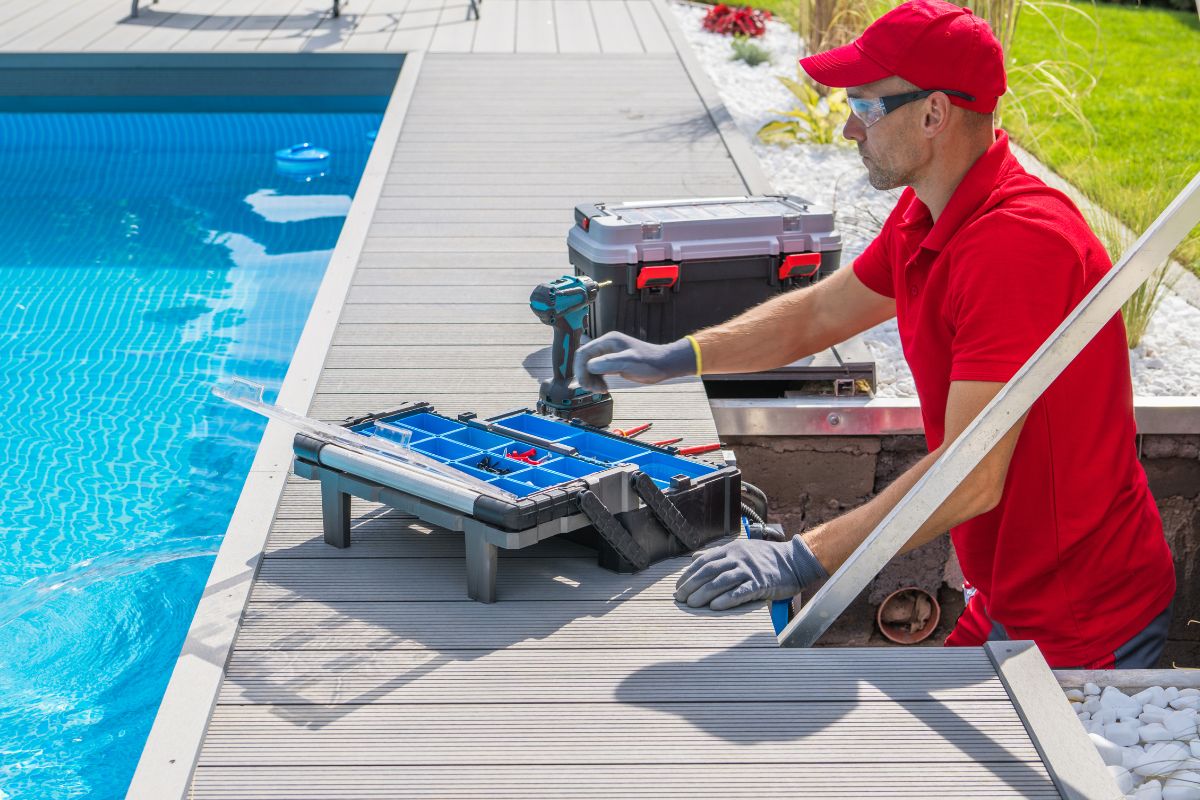 A worker in red uniform, gloves, and safety glasses performs seasonal pool maintenance, repairing equipment beside an outdoor swimming pool, with tools and a toolbox on the pool deck.