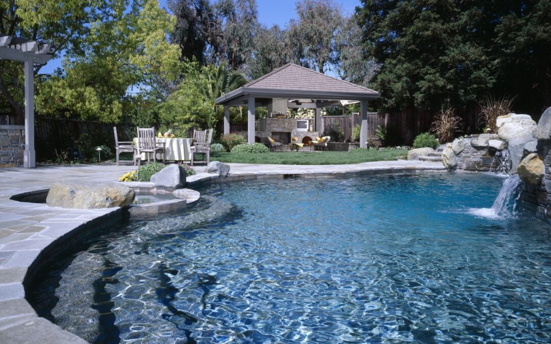 A backyard featuring a clear inground swimming pool with a waterfall, surrounded by rocks. In the background, there's a gazebo, a green lawn, and a wooden outdoor seating area under a pergola with trees around. The water features add an element of tranquility to the space.