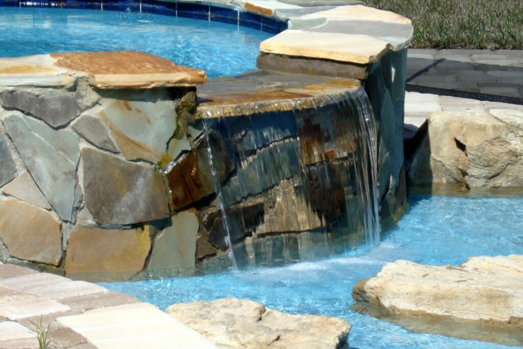 A stone waterfall feature flows into an inground pool, with blue water cascading over the edge into a shallow area surrounded by rocks.