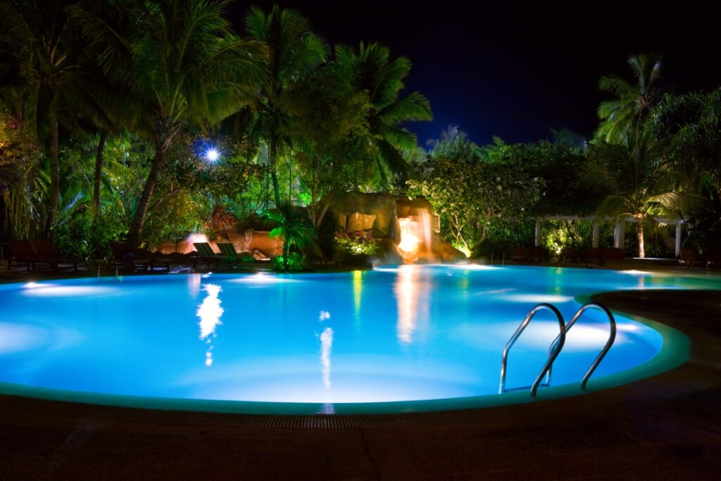 A brightly lit inground swimming pool at night, surrounded by palm trees and greenery, features a metal ladder extending into the water and captivating water features, including a waterfall visible in the background.