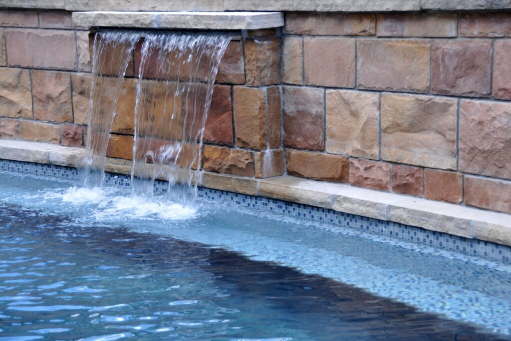 Water cascades from a stone structure into an inground pool, creating elegant water features that complement the tiled edge and brick wall backdrop.