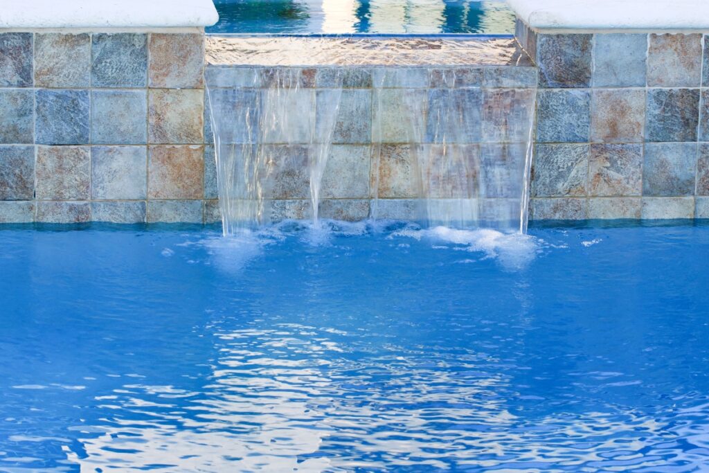 Water flows from a tiled fountain feature into an inground pool, creating a cascading effect. The pool water is clear and blue, showcasing one of the finest inground pool water features.