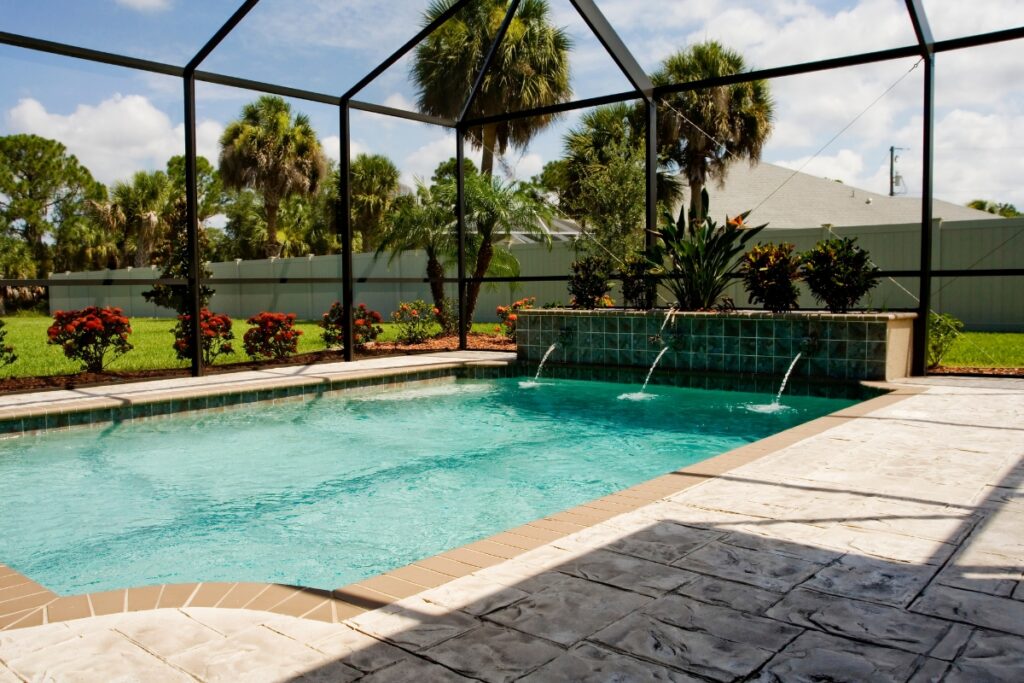 A backyard pool area featuring a screened enclosure, inground pool water features with a small waterfall, and surrounding landscaping with palm trees and shrubs.