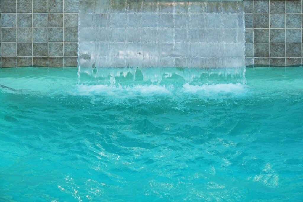 A clear waterfall flows from a tiled wall into an inground pool, creating small splashes and ripples on the turquoise water surface, enhancing the luxury of backyard water features.