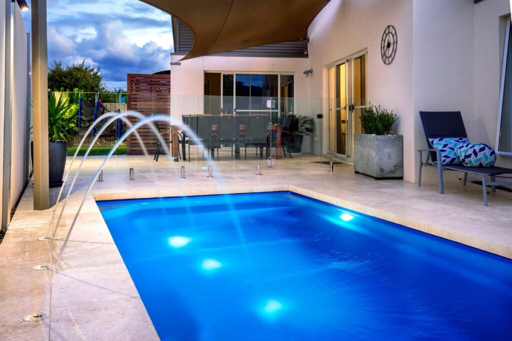 A modern backyard features a small rectangular inground pool with water jets and lights, surrounded by a tiled patio. There's outdoor furniture, a dining table, and a covered seating area against the house.