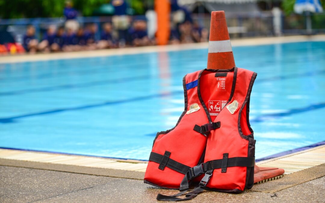 A red life vest and an orange traffic cone, placed for pool safety regulations, on the edge of a swimming pool, with blurred swimmers in the background.