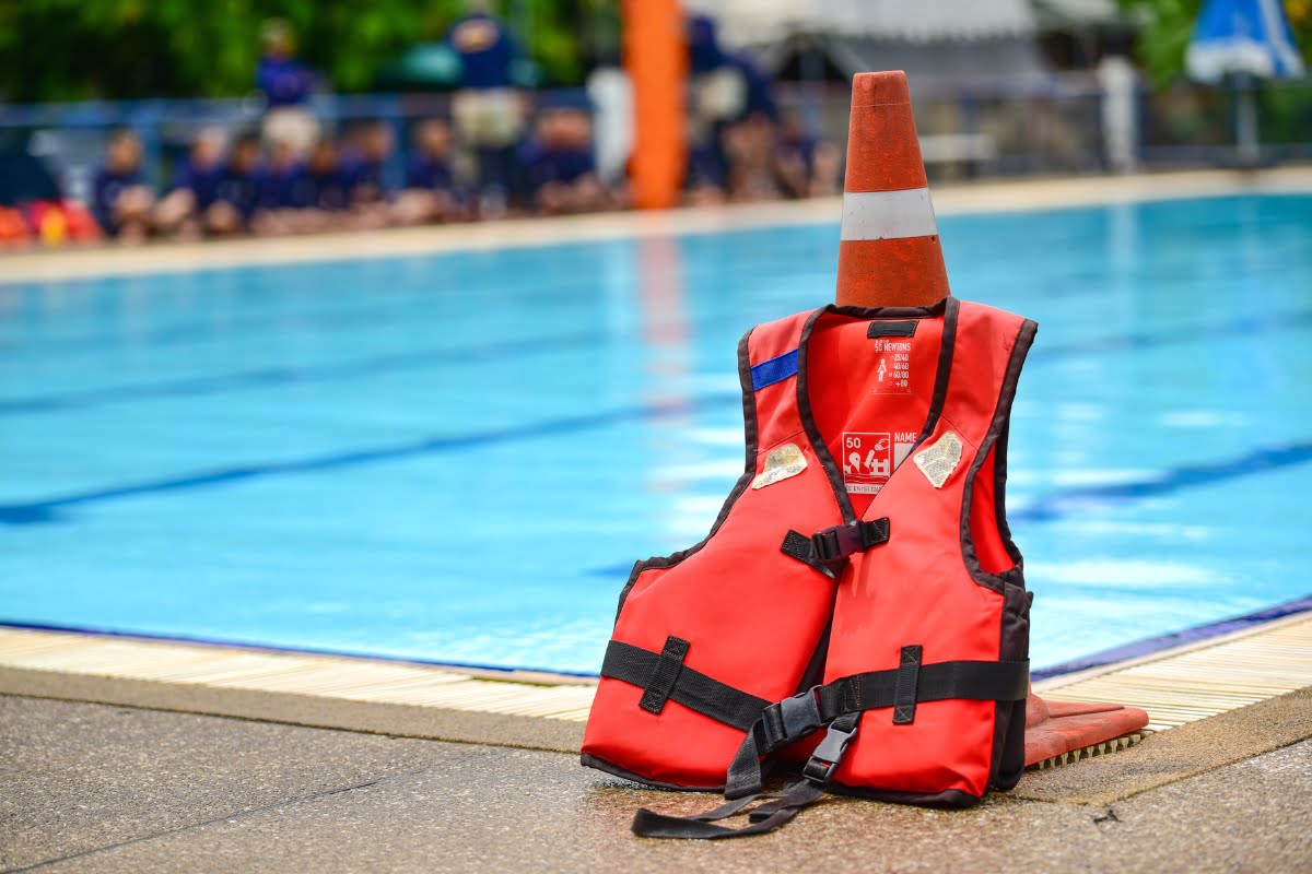 A red life jacket and an orange traffic cone are placed on the poolside next to a swimming pool, following pool safety regulations, with people blurred in the background.