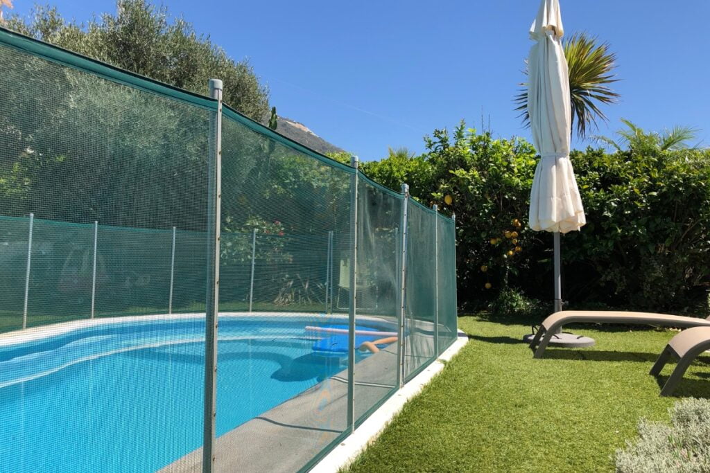 A residential outdoor swimming pool compliant with pool safety regulations, enclosed by a transparent safety fence, with sun loungers and a closed white umbrella on a grassy lawn, under a clear blue sky.