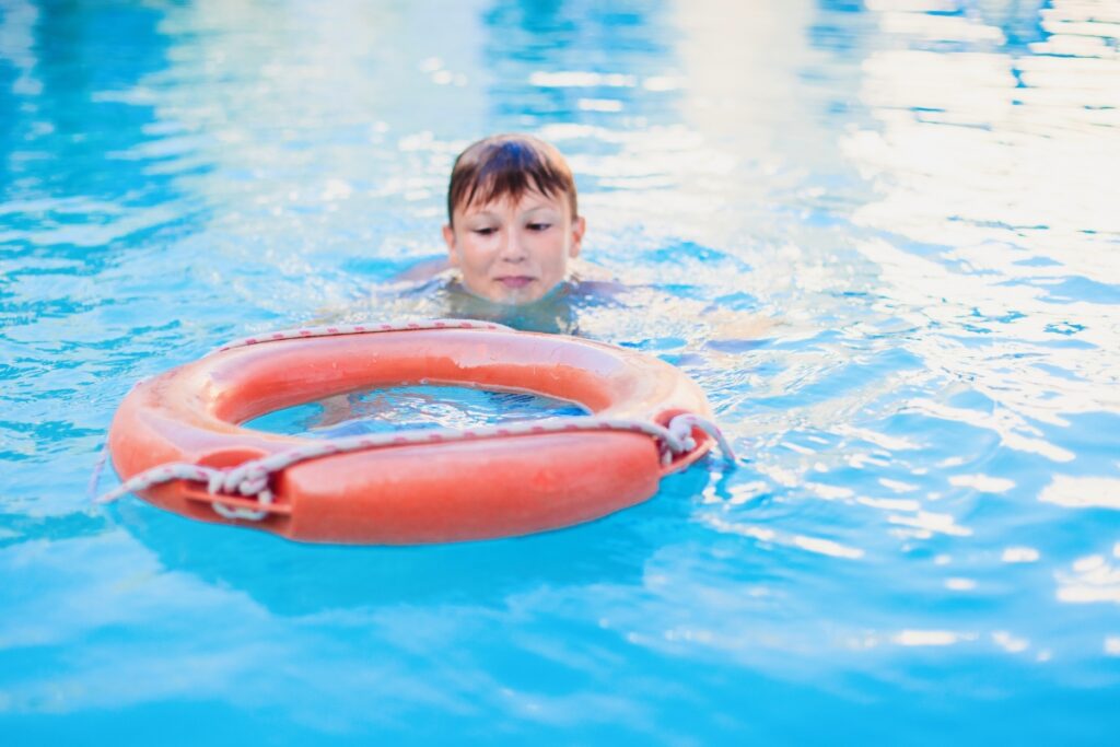 Young boy swimming towards an orange lifebuoy in a clear blue pool, adhering to pool safety regulations.