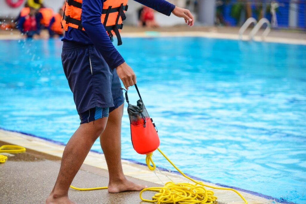 A lifeguard in blue shorts and a life vest adheres to pool safety regulations while performing equipment maintenance beside a swimming pool, holding a rescue buoy and rope.