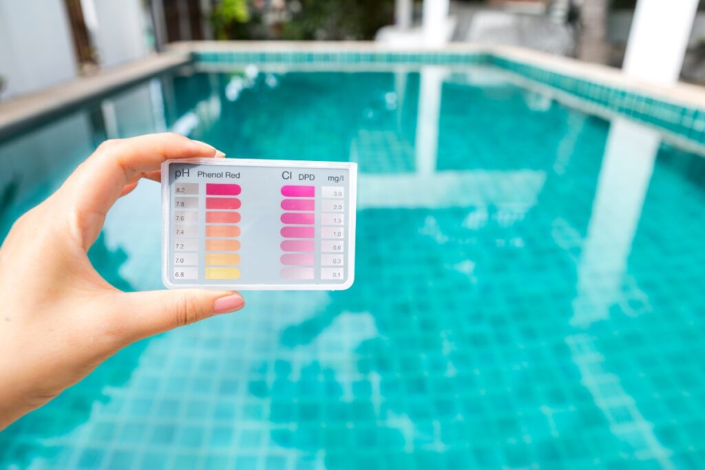 A hand holding a pH test strip chart in front of a clear, turquoise swimming pool for water quality testing in compliance with pool safety regulations.