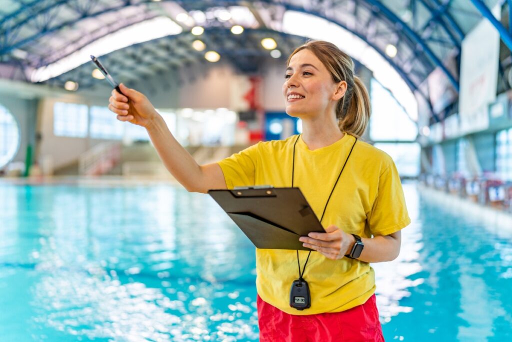 A female coach in a yellow shirt and red shorts smiling and taking a selfie with a smartphone by an indoor swimming pool, overseeing pool safety regulations.