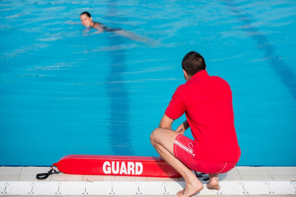 Lifeguard monitoring pool safety regulations in a red uniform, sitting at the edge of a pool, watching a swimmer in the water.