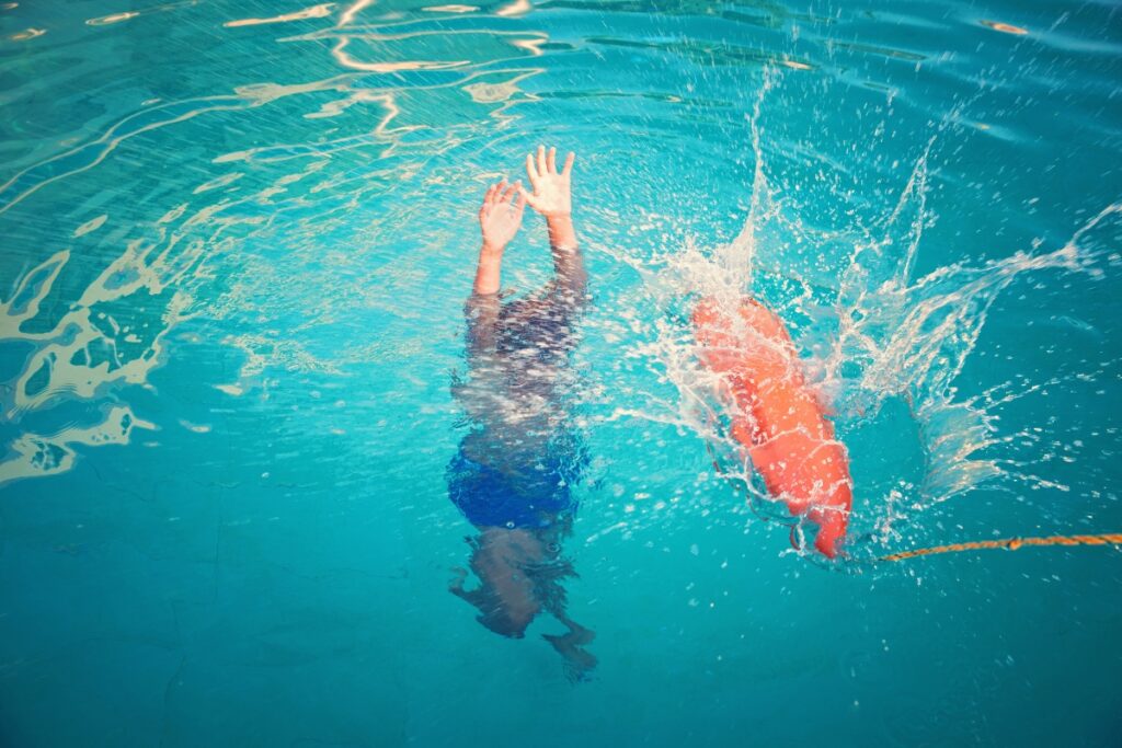 A child in a blue and black swimsuit is submerged, reaching towards the surface of a vibrant teal swimming pool, adhering to pool safety regulations, creating splashes.