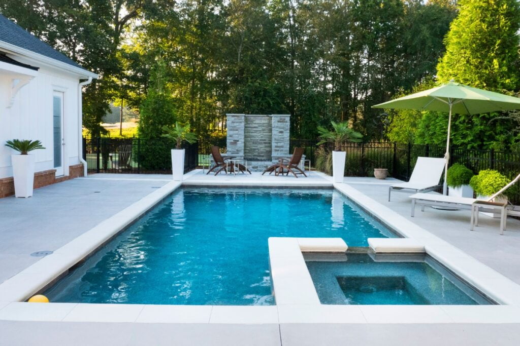 A modern outdoor swimming pool area featuring stylish outdoor poolside decor, lounge chairs, a green umbrella, and a small hot tub, all surrounded by lush trees and greenery.