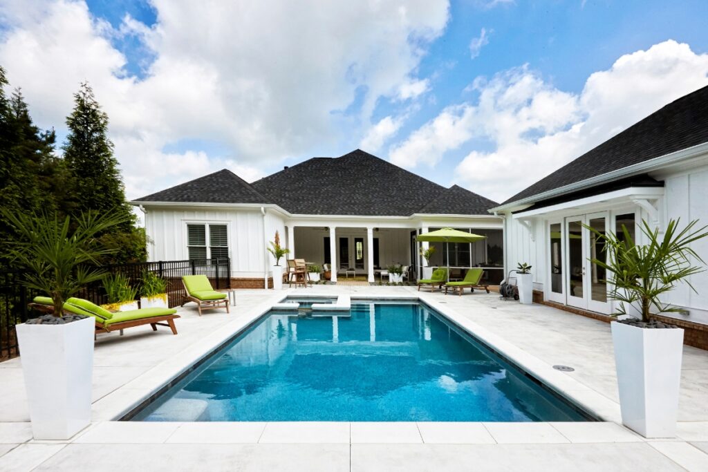 A backyard view features a rectangular swimming pool surrounded by lounge chairs and outdoor poolside decor, including large white planters with plants. A light-colored house with patios and windows complements the scene under a partly cloudy sky.