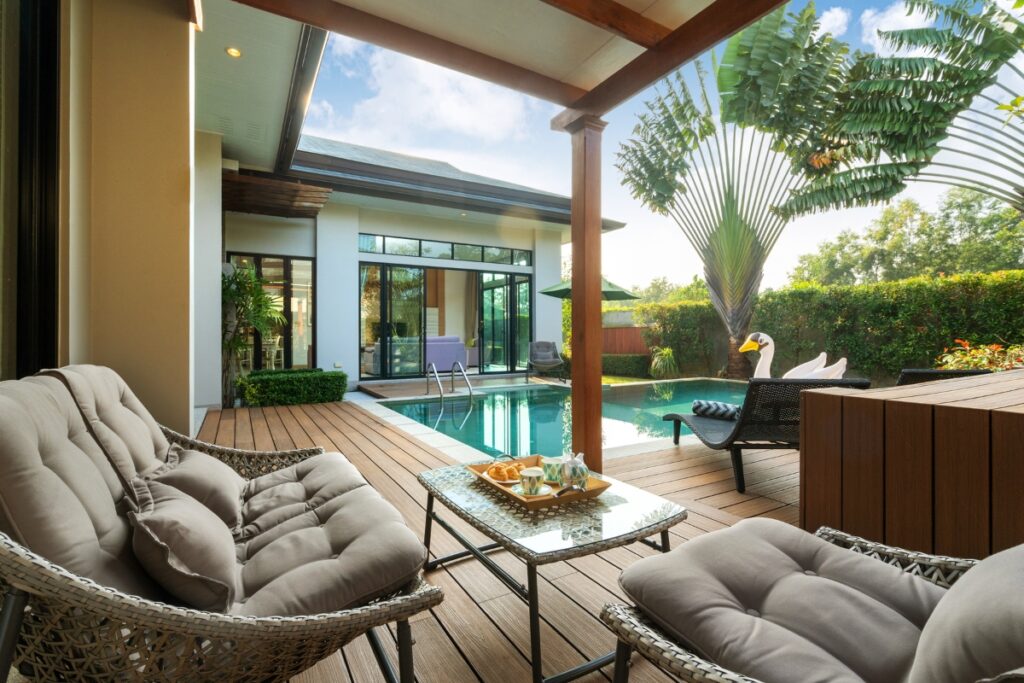 Luxurious outdoor patio with cushioned seating, a glass-top table with snacks, and a view of a swimming pool and modern house. The outdoor poolside decor is complemented by lush greenery that surrounds the area.