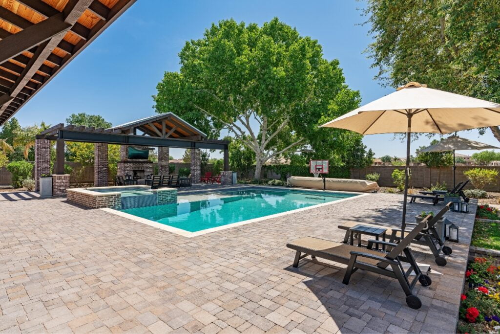 Outdoor pool area with elegant poolside decor, lounge chairs, a large umbrella, and a covered patio with a fireplace. Paved surroundings and trees in the background under a clear blue sky create an inviting atmosphere.