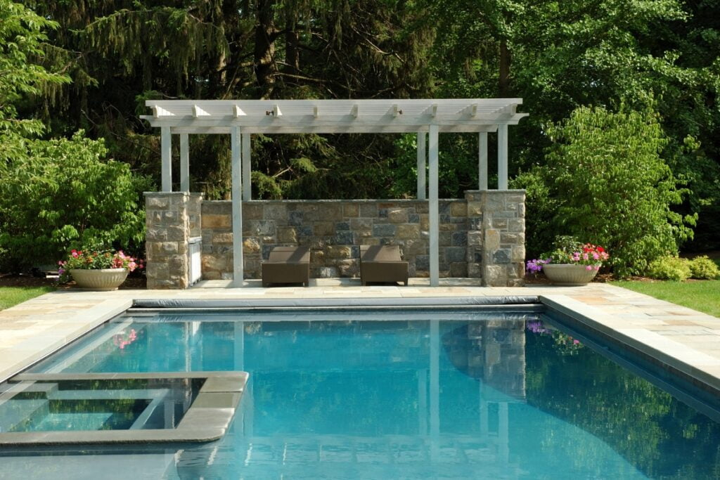 A rectangular swimming pool with clear blue water, surrounded by greenery and trees, features a pergola with two lounge chairs and tasteful outdoor poolside decor against a stone wall backdrop.