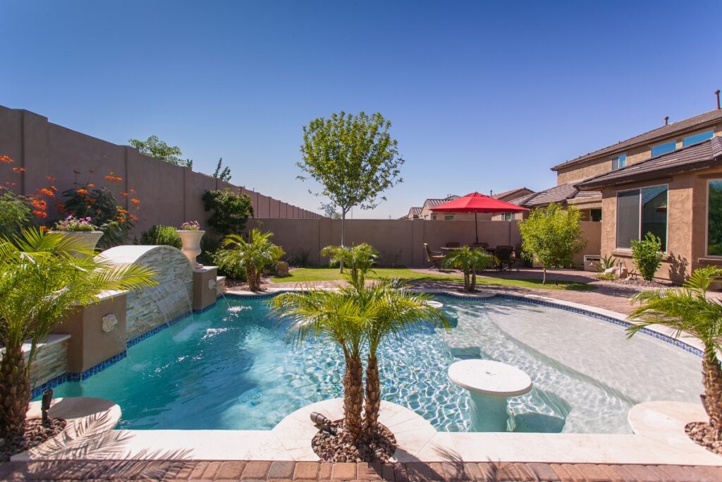 A backyard features a swimming pool with palm trees, a water feature, stylish outdoor poolside decor, and a patio with a red umbrella. The well-maintained landscaping adds charm, while a wooden privacy fence encloses the area.