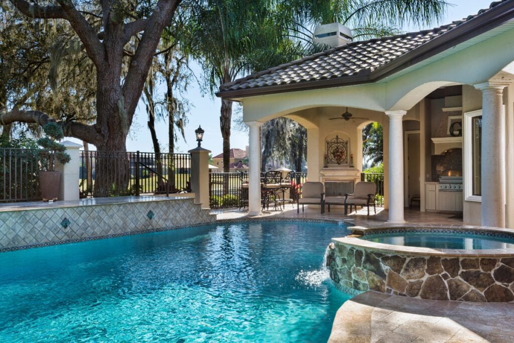 A luxurious backyard features a swimming pool, hot tub, and tasteful outdoor poolside decor, along with a patio area with seating and a large tree providing shade, next to a house with an open columned porch.