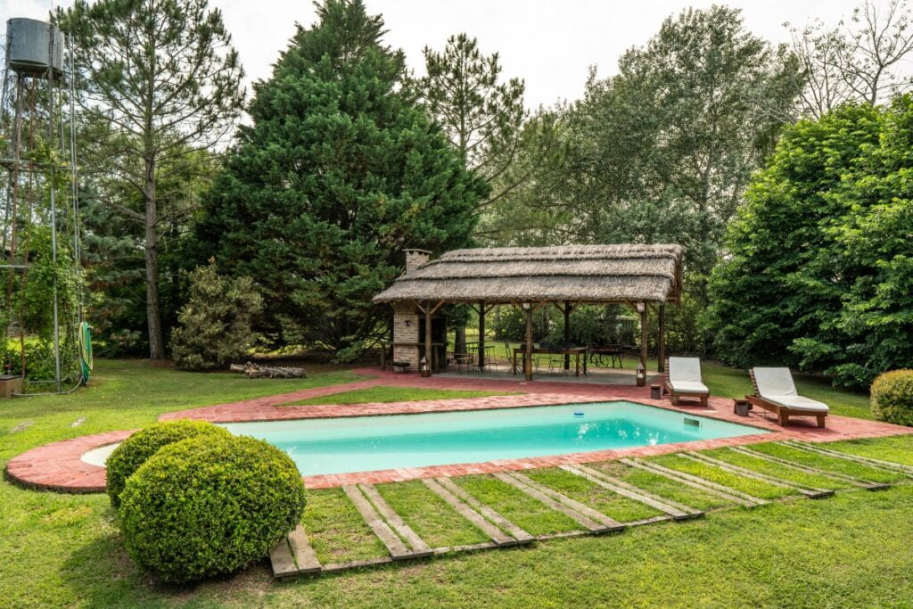 A backyard features a rectangular swimming pool surrounded by grass, two lounge chairs on a brick path, and a thatched-roof pavilion with an outdoor dining area, all framed by lush green trees and shrubs to create perfect outdoor poolside decor.