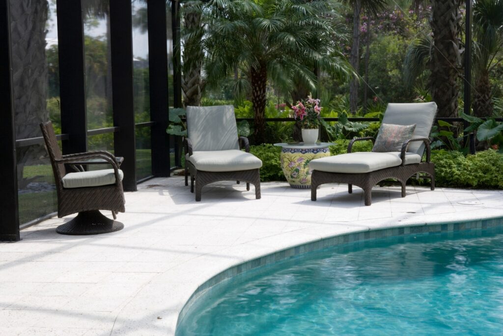 Outdoor patio featuring three wicker chairs around a small table with a floral arrangement, all as part of the elegant outdoor poolside decor, next to a swimming pool surrounded by lush greenery.