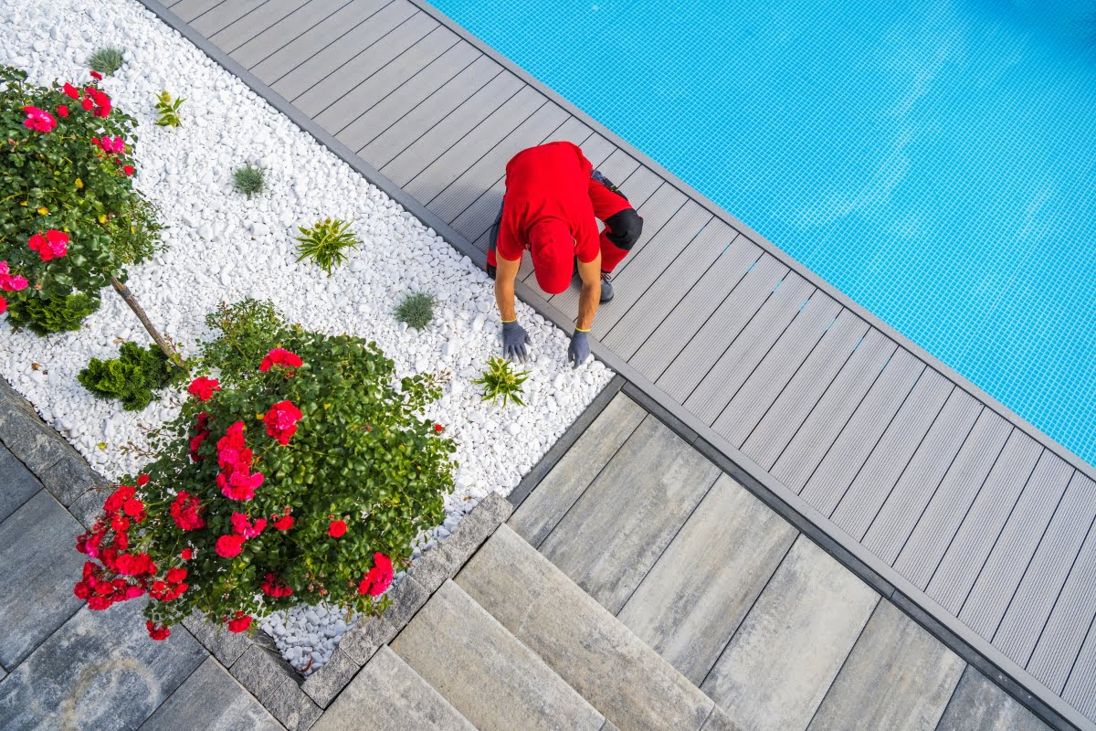 Person in red shirt and cap engaged in poolside gardening, tending to plants in a landscaped garden next to a swimming pool, with stone and wooden decking visible.