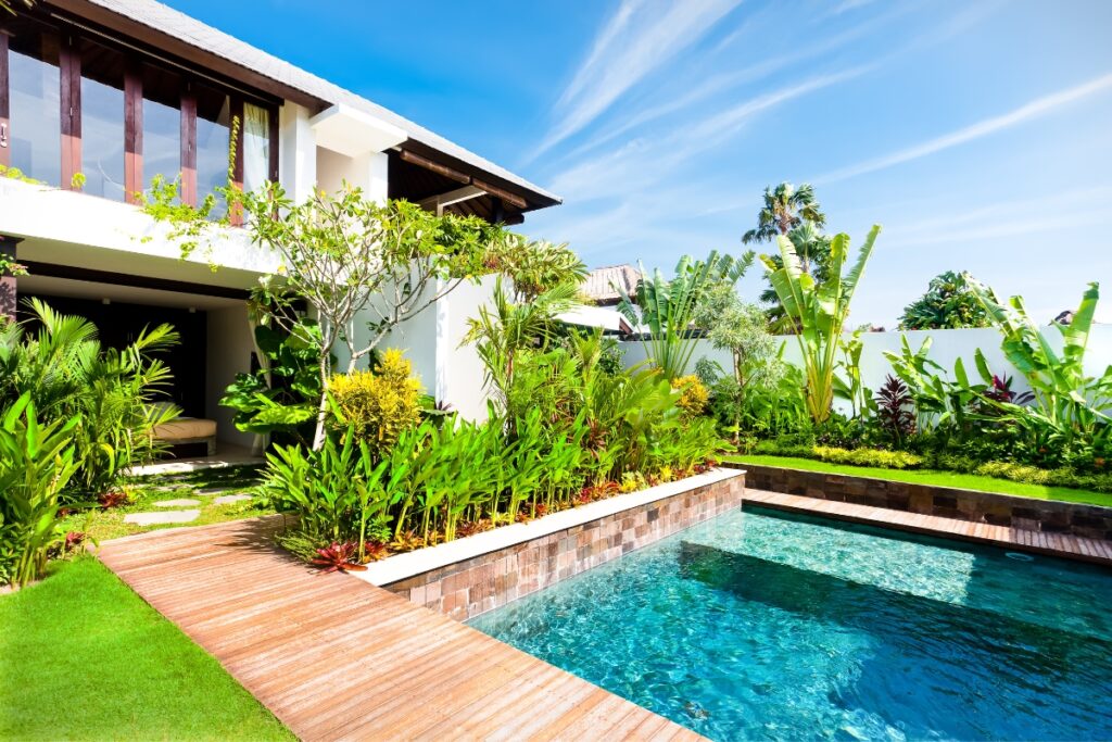 A modern two-story house with a manicured garden, wooden deck, and a rectangular swimming pool under a bright blue sky offers the perfect setting for poolside gardening enthusiasts.