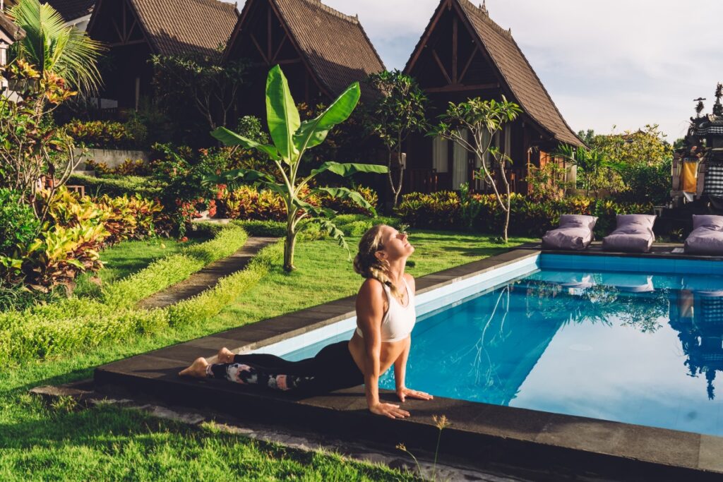 A person practices yoga beside a swimming pool in a lush garden, engaging in some poolside gardening, with traditional-style buildings in the background.