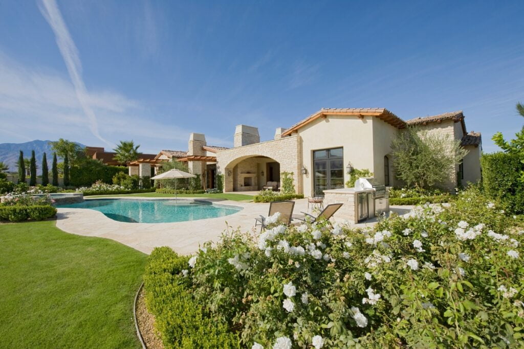 A spacious villa with a red-tiled roof features a swimming pool, lounge chairs, and a beautifully landscaped garden with white flowers and shrubs perfect for poolside gardening, all set against a backdrop of mountains under a clear blue sky.