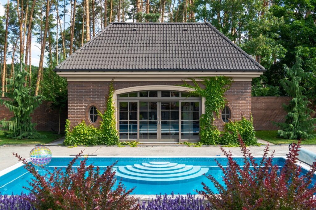 A small brick pool house with a gabled roof stands behind a round swimming pool with concentric steps, surrounded by lush greenery and a garden featuring blooming shrubs and trees, perfect for poolside gardening.