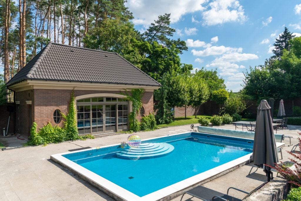 A contemporary backyard features a rectangular swimming pool with a circular step entry, adjacent spa, ivy-covered brick guesthouse, umbrella, lounge chairs, and surrounding trees under a blue sky—perfect for poolside gardening.