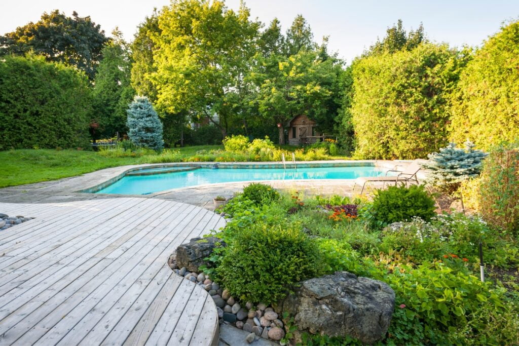 A backyard scene with a wooden deck, landscaped garden, and a sparkling swimming pool. Trees and shrubs surround the pool area, perfect for poolside gardening enthusiasts. There's also a small structure or shed in the background, adding to the charm of this serene setting.