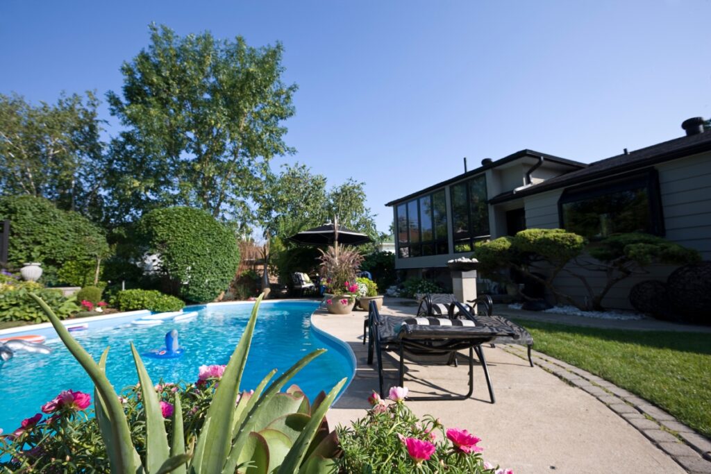 A backyard with a swimming pool, lounge chairs, umbrella, and various blooming flowers showcasing poolside gardening in the foreground. A modern house stands in the background with large windows and trees surrounding the area.