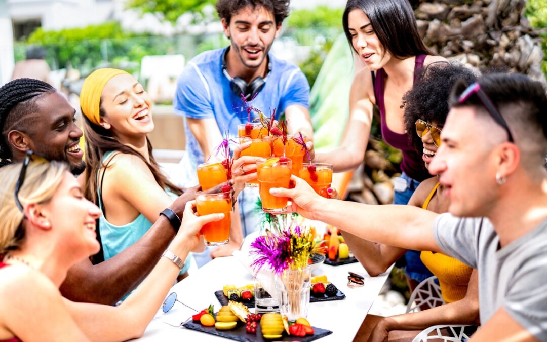 A group of eight people clink glasses while seated around a table with food and drinks outdoors, perfect for gathering poolside party ideas.