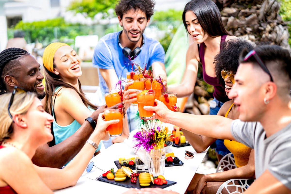A group of people sit around an outdoor table, smiling and raising glasses filled with orange drinks, with food and flowers on the table—perfect inspiration for poolside party ideas.