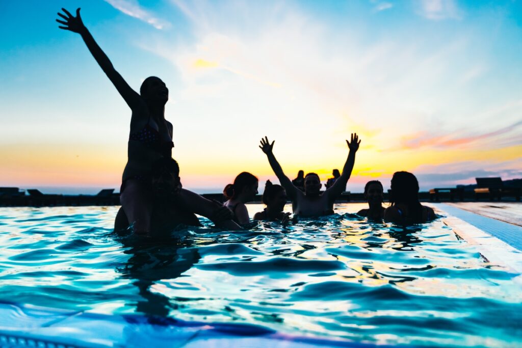 A group of people enjoying themselves in a swimming pool at sunset, with some raising their arms in delight, showcases perfect poolside party ideas.