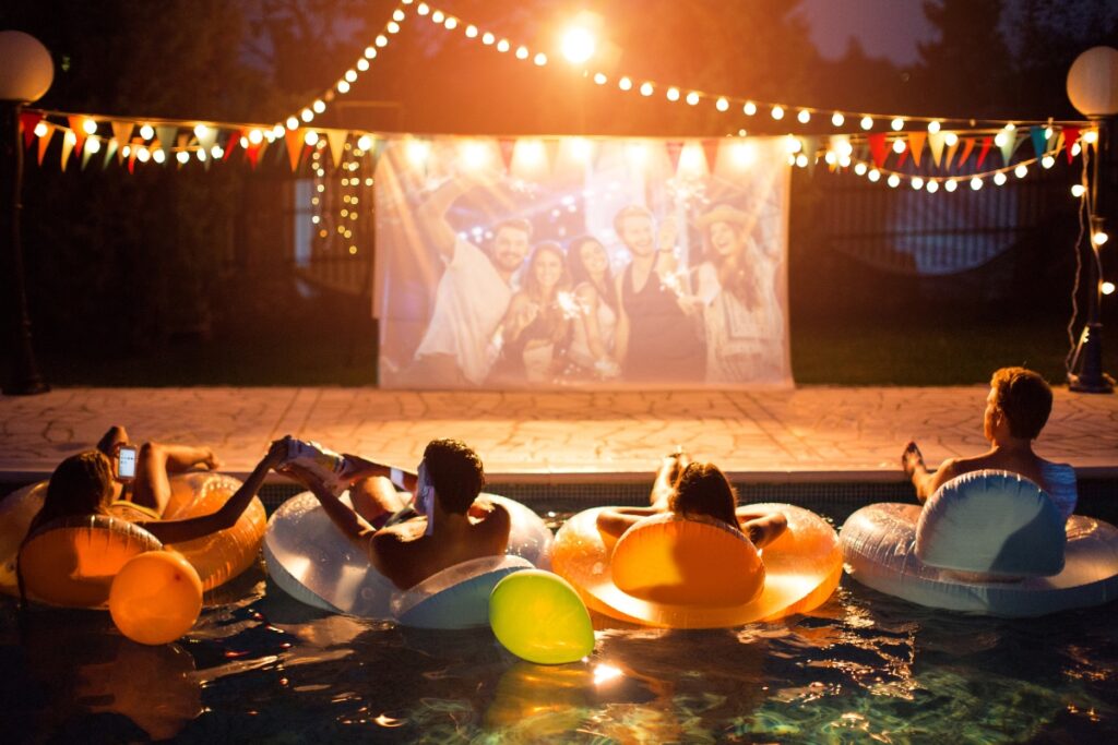 Four people float on inflatable loungers in a pool at night, enjoying a poolside party with a movie projected on a screen, string lights, and festive flags overhead.