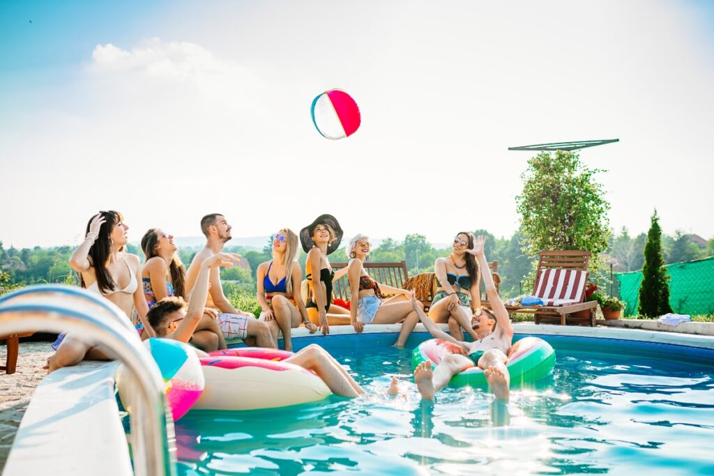 A group of people in swimsuits enjoy a sunny day by playing with a beach ball in and around an outdoor pool, showcasing perfect poolside party ideas.