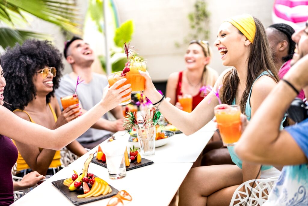 A group of people sitting at a table outside, smiling, and raising colorful drinks with fruit garnishes. Plates with fruit arrangements are on the table, offering perfect poolside party ideas.