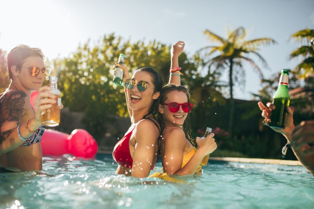 Three people in a pool holding drinks and wearing sunglasses on a sunny day with palm trees in the background, offering perfect poolside party ideas.