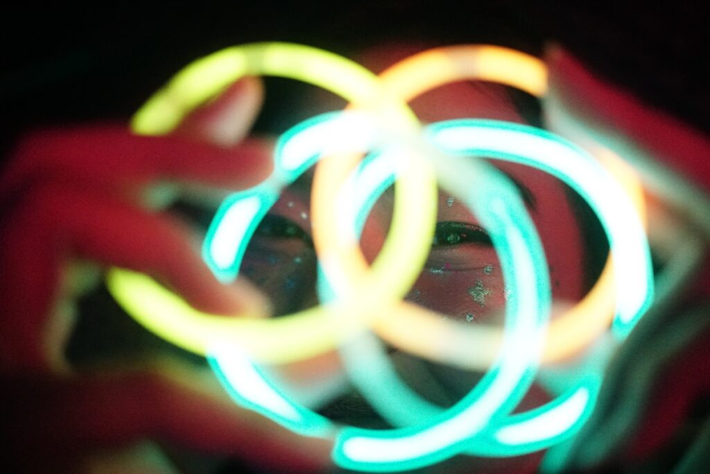 A person with face paint holds colorful, glowing rings close to their face, creating a vibrant and illuminated scene perfect for your next poolside party.