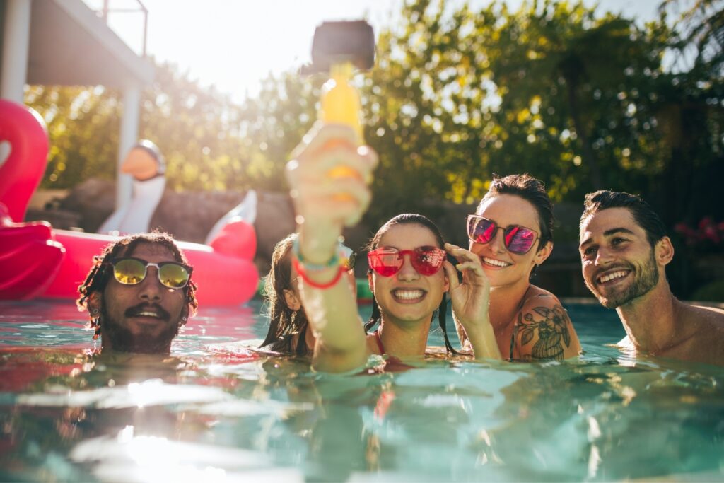 Four people in a pool wearing colorful sunglasses and holding a selfie stick celebrate one of the best poolside party ideas, with inflatable floats adding to the fun atmosphere in the background.