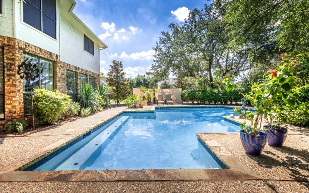 A backyard with a swimming pool, surrounded by greenery and potted plants, offers serene privacy solutions. A two-story house stands on the left with windows overlooking the pool area. Clear blue sky above.