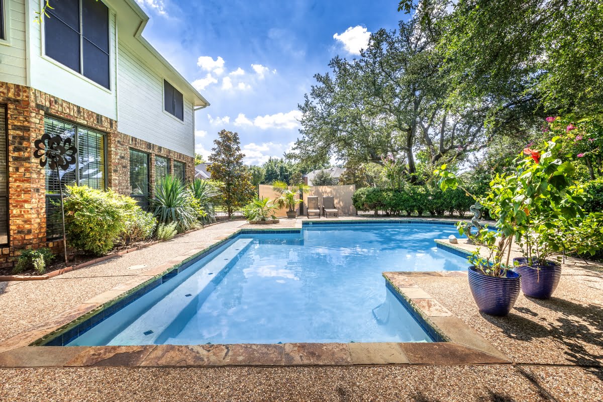 A backyard with a rectangular swimming pool and spa, surrounded by plants and trees that offer natural privacy solutions, next to a brick and white two-story house under a sunny sky.