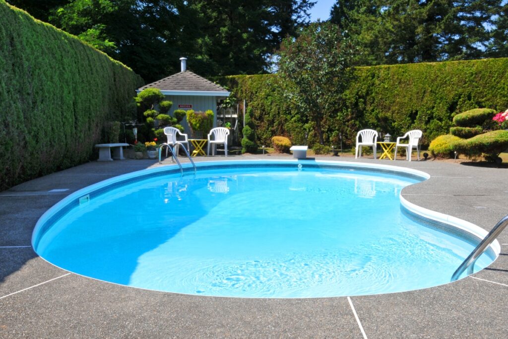 A clean, curvy outdoor swimming pool is surrounded by a well-maintained hedge offering privacy solutions, white chairs, and a small outbuilding, all bathed in sunlight.