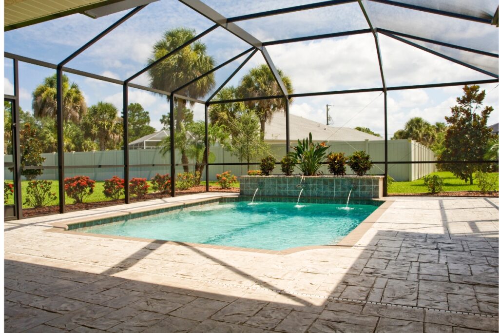 A backyard swimming pool with an adjacent waterfall feature, enclosed by a screened-in structure for added privacy solutions. The area includes well-kept landscaping with palm trees and various plants under a clear sky.